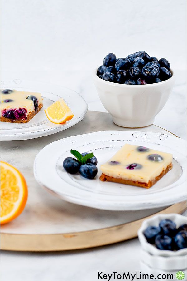 A side image of two lemon blueberry bars on white plates on top of a marble slab with a bowl of blueberries in the background.