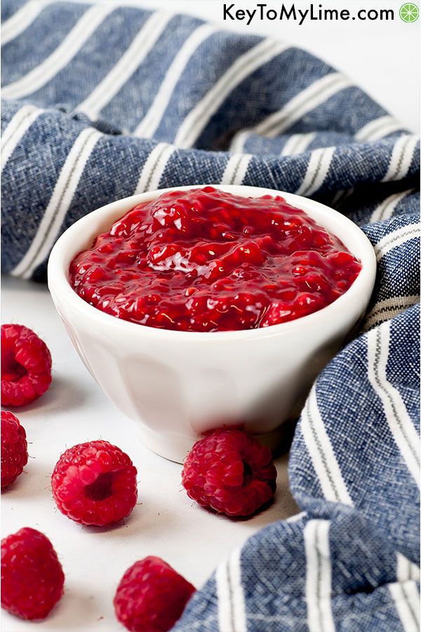 Raspberry sauce in a small white bowl surrounded by raspberries and a blue napkin.