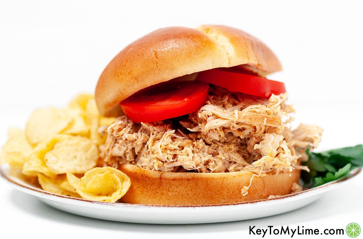 A pulled chicken sandwich next to potato chips against a white background.