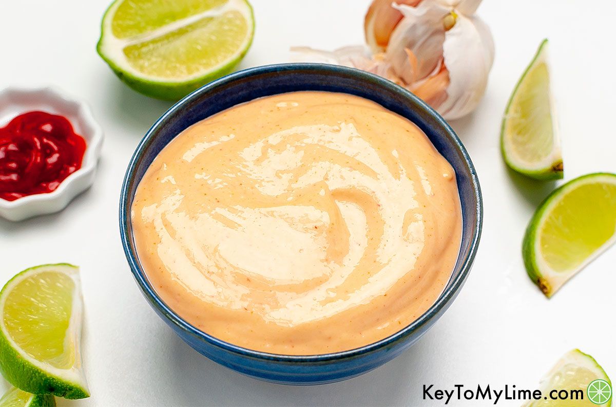 A small bowl of sriracha mayo surrounded by garlic and lime wedges.
