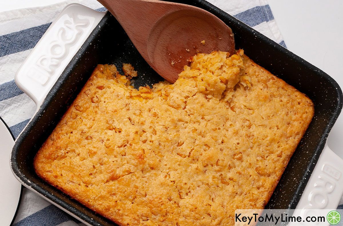 A large wooden spoon scooping corn casserole out of the baking dish.