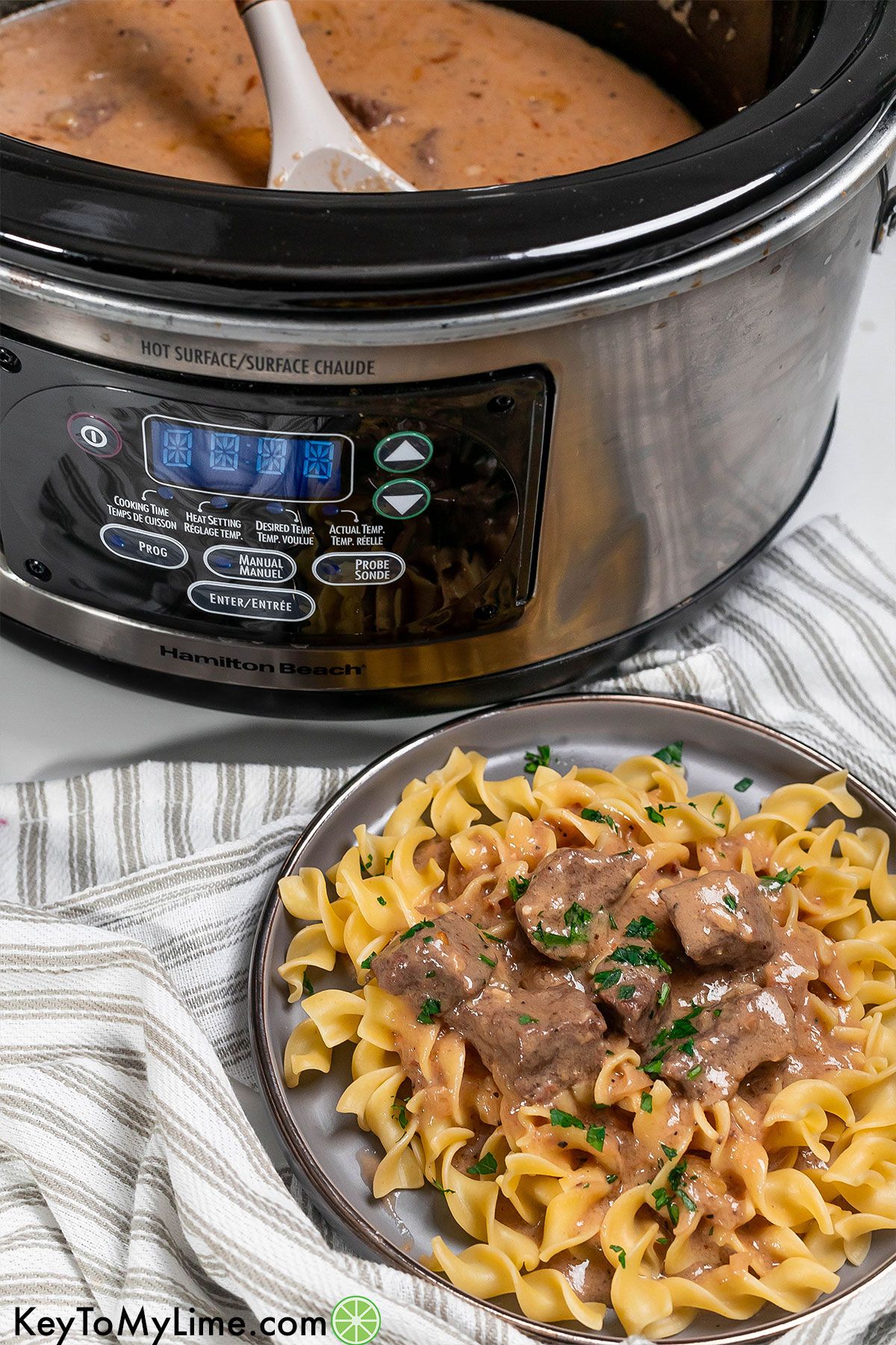 Crockpot beef tips plated on egg noodles with a crockpot in the background.