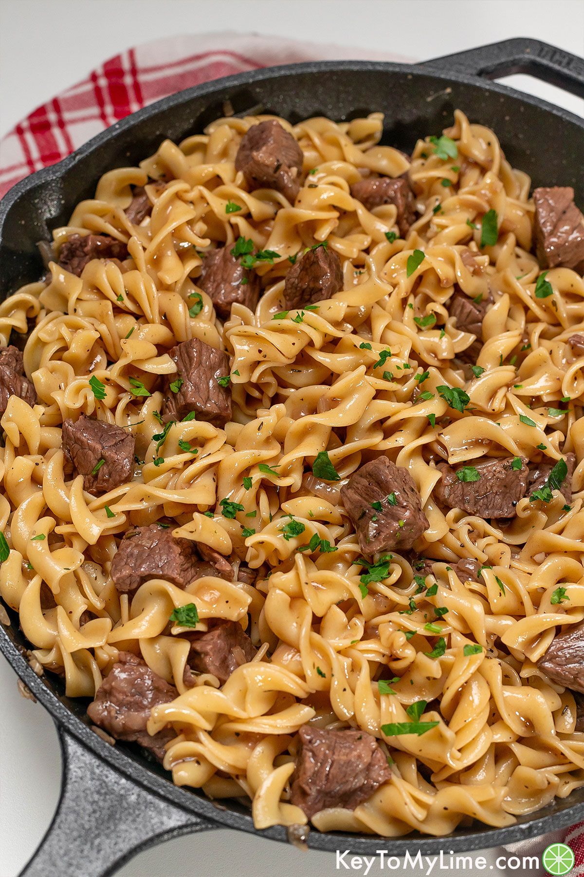 Tender and savory beef tips and noodles served in a large iron skillet with fresh parsley throughout.