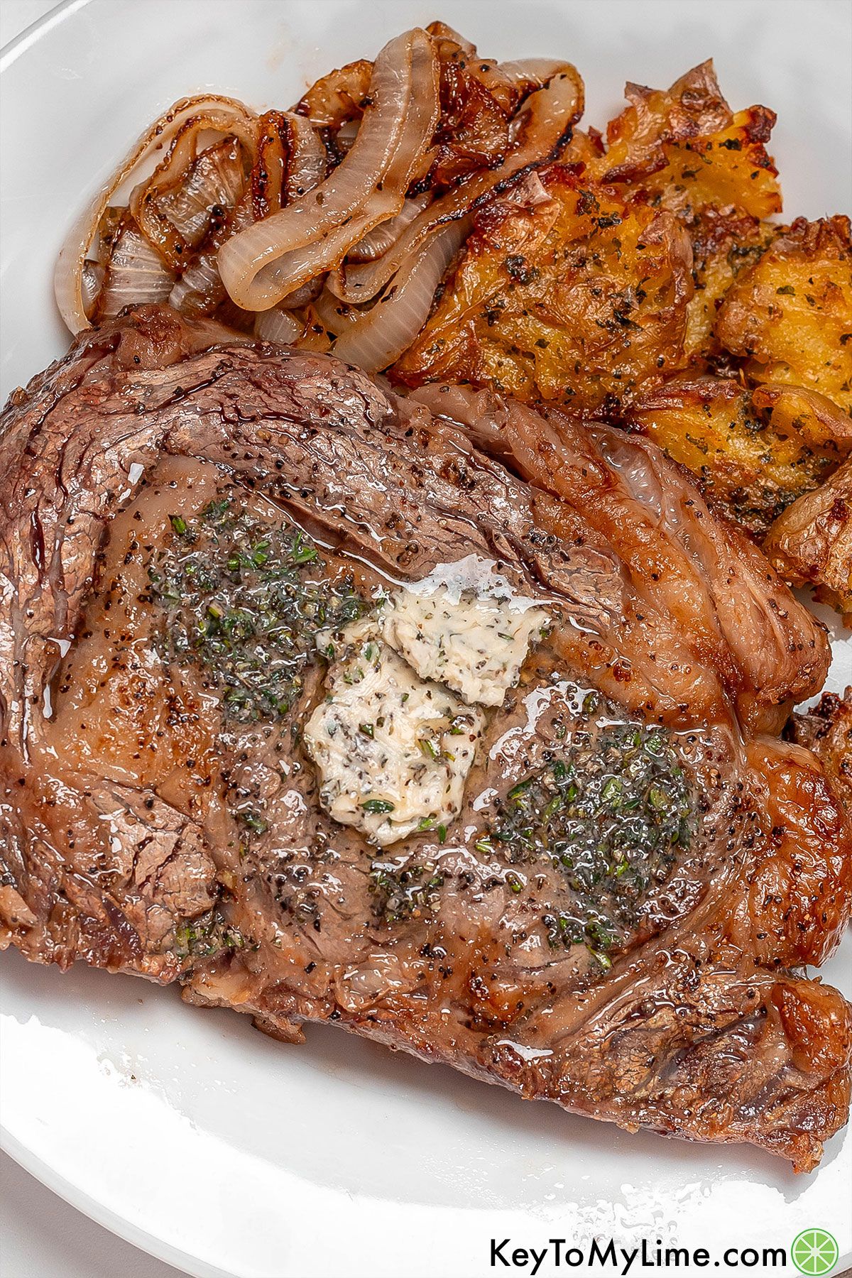 An overhead image of a cooked ribeye with glistening herb butter throughout served a white plate.